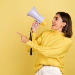 young-woman-yellow-warm-sweater-with-megaphone-speaker-screaming-left-pointing-index-finger