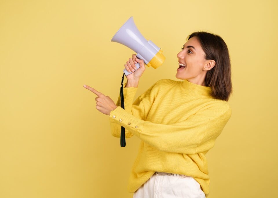 young-woman-yellow-warm-sweater-with-megaphone-speaker-screaming-left-pointing-index-finger